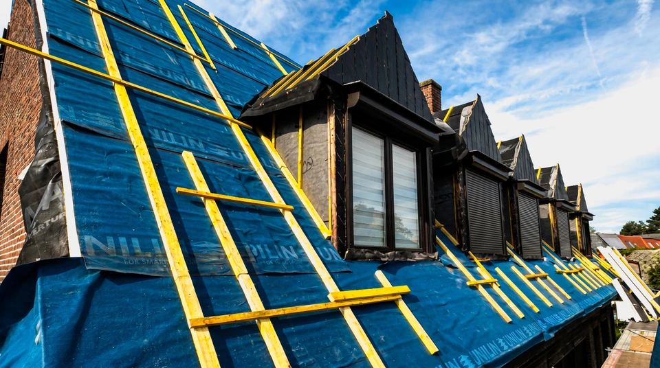 Photographie r&eacute;aliste en ext&eacute;rieur montrant un couvreur exp&eacute;riment&eacute; travaillant sur une toiture de maison r&eacute;sidentielle classique. L'artisan, v&ecirc;tu de tenue professionnelle appropri&eacute;e et &eacute;quipement de s&eacute;curit&eacute;, effectue des travaux de couverture dans un environnement authentique. La perspective et la composition mettent en avant l'engagement professionnel et le savoir-faire artisanal du travail de couverture, avec visibilit&eacute; claire du chantier r&eacute;el et de la concentration du professionnel &agrave; la t&acirc;che. L'arri&egrave;re-plan montre l'environnement r&eacute;sidentiel pertinent &agrave; l'activit&eacute; locale de l'entreprise aupr&egrave;s des particuliers.